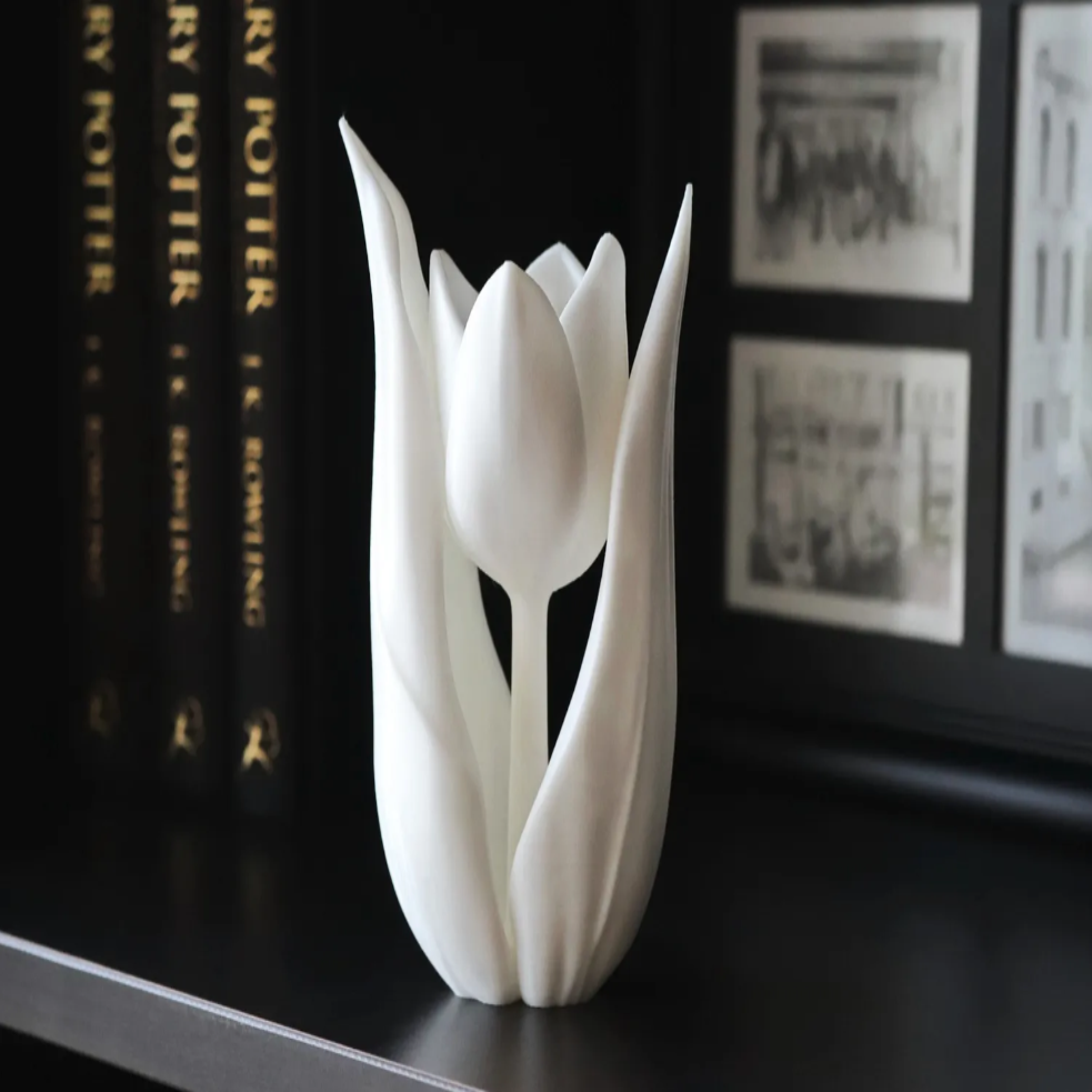 A 3D printed white flower decoration placed on a shelf with books in the background.