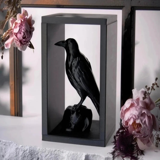 Black crow figurine in a shadow box with pink flowers on a white surface